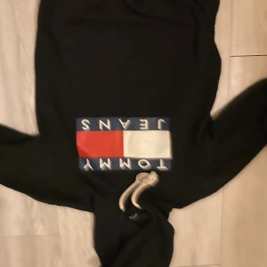  Tommy Jeans hoodie  - Svart hoodie från Tommy Jeans med stor klassisk logga i rött, vitt och blått framtill. Tröjan har huva med vita snören och en stor magficka. Perfekt för dig som gillar streetwear och vill ha en stilren look.