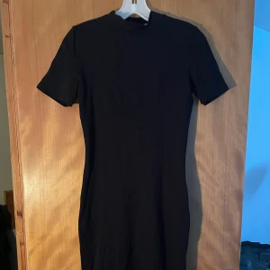 Svart kortärmad midiklänning  - En enkel och stilren svart klänning med korta ärmar och rund hals. Modellen är figurnära och har en kort längd som ger en modern vibe. Perfekt för dig som gillar minimalistisk stil och vill ha en klänning som är lätt att matcha med accessoarer.