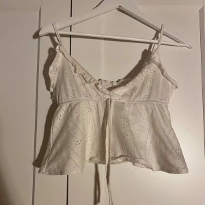 Vit linnetopp med snörning - Säljer en söt vit linnetopp med broderade detaljer och smala axelband. Toppen har snörning framtill och en lätt volangkant vid axlarna. Perfekt för varma dagar och ger en romantisk vibe till din outfit.