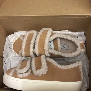 Beige sneakers från Inuikii med fuskpäls - Unika beige sneakers från Inuikii med fluffiga detaljer i fuskpäls och mocka. Skorna har platt vit sula och breda kardborreband med päls runtom. Perfekt för dig som gillar cozy vibes och vill sticka ut med en cool look. Fick dessa i födelsedags present men tyvärr så var de för stora:(