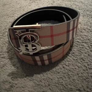 Burberry-bälte  - Snyggt bälte från Burberry med klassiskt rutigt mönster i beige, rött, svart och vitt. Bältet har ett stilrent spänne i silver med Burberrys ikoniska 'B'-logga. Perfekt accessoar för att lyfta din outfit med en lyxig touch.