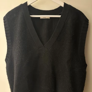 Svart stickad väst från Gina Tricot - Svart stickad väst från Gina Tricot med djup v-ringning och breda ribbade kanter vid ärmhålen och nederkant. Västen har en enkel och clean look som är lätt att styla över en skjorta eller t-shirt. Perfekt för lager-på-lager under hösten och vintern.