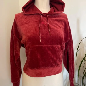 Vinröd velour hoodie från Puma - Säljer en vinröd hoodie från Puma i mjuk velour med croppad passform. Tröjan har huva med snörning, stor magficka och långa ärmar. Perfekt för dig som gillar sportig och chill stil.