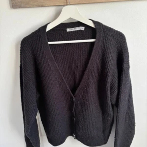 Svart stickad kofta från NA-KD - Mysig svart stickad kofta från NA-KD i storlek XL. Koftan har v-ringning, knappar framtill och långa ärmar. Perfekt för lager-på-lager och enkel att matcha med olika outfits. Finns lite ytludd.
