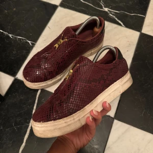 Vinröda sneakers med reptilmönster - Säljer ett par vinröda sneakers från Giuseppe Zanotti med reptilmönstrad yta i skinn och snygga guldfärgade dragkedjor på ovansidan. Skorna har platt vit sula och rund tå, vilket ger en cool och lyxig vibe. Perfekt för dig som gillar statement pieces.
