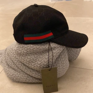 Svart keps från Gucci med GG-mönster - Säljer en svart keps från Gucci med diskret GG-monogram över hela kepsen. Klassisk böjd skärm och justerbart spänne bak i skinn. Sidorna har Guccis ikoniska gröna och röda band. Perfekt accessoar för dig som vill sticka ut med lyxig streetstyle.
