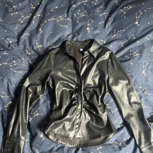 Svart skjorta i skinnimitation med knappar - Svart långärmad skjorta från Vinted i skinnimitation med snygg rynkning längs knappraden framtill. Skjortan har klassisk krage, figurnära passform och blanka knappar. Perfekt för dig som gillar en snygg fest topp.