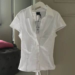 Vit blus med knyt från Gina Tricot - Snygg vit blus från Gina Tricot i storlek XS/S. Modellen har korta ärmar, klassisk krage och knappar framtill. Blusen är figursydd med knytband i midjan för extra form. Aldrig använd, prislappen finns kvar!
