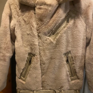 Beige pälsjacka med bälte och dragkedja - Mysig och fluffig beige pälsjacka med bred krage, snedställd dragkedja och bälte i midjan. Jackan har två fickor med dragkedja och en oversized känsla. Perfekt för kyliga dagar och ger en trendig look.