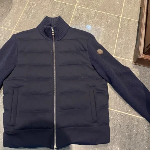 Mörkblå Moncler cardigan tricot - Snygg mörkblå cardigan från Moncler med quiltad front och stickade ärmar. Jackan har hög ribbad krage, dragkedja med läderdetalj och Moncler-logga på ärmen. Bra skick och väldigt fin jacka. Även äkta! 