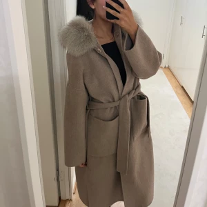 Beige kappa med pälsdetalj och bälte - Snygg beige kappa med stora fickor, bälte i midjan och fluffig ljusgrå pälsdetalj på kragen. Storlek M men skulle säga den är Onesize. Fint skick. Lite nopprig på vissa ställen men inget man lägger märke till. 