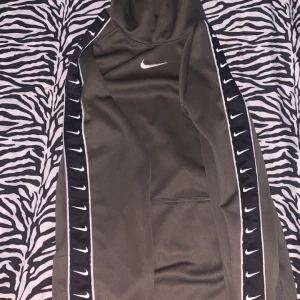 Nike hoodie med loggor på ärmarna - Nike hoodie i svart med vita detaljer och Nike-loggor längs båda ärmarna. Tröjan har huva, lång ärm och en klassisk sportig look. Materialet är mjukt och känns som polyester, perfekt för chill eller träning.