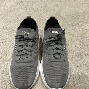 Grå sneakers från Björn Borg - Snygga grå sneakers från Björn Borg med vit sula och stickad ovandel i syntetmaterial. Skorna har snörning och en modern, sportig design med rund tå och platt sula. Perfekta för dig som gillar stilrena och bekväma sneakers med diskret logga på sidan. Skorna har använts en gång.