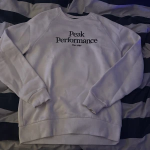 Vit sweatshirt från Peak Performance - Vit sweatshirt från Peak Performance med svart broderad logga på bröstet. Tröjan har rund hals, ribbade muddar och är långärmad. Mjuk insida och klassisk passform, perfekt för en clean och sportig look.