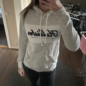 Ljusgrå hoodie från Hollister  - Säljer en ljusgrå hoodie från Hollister i storlek XS. Tröjan har broderad logga framtill, snörning i huvan och en stor magficka. 