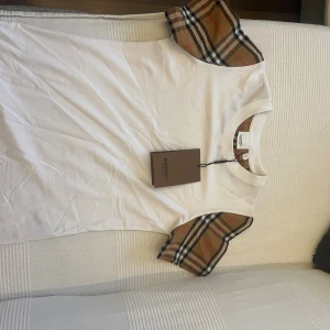 Vit Burberry t-shirt med rutiga ärmar - Säljer en fräsch vit t-shirt från Burberry med klassiska rutiga detaljer på ärmarna och runt halsen. T-shirten har korta ärmar och är gjord i mjuk bomull. Perfekt för dig som gillar stilrena plagg med lyxig touch.