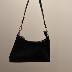 Svart axelväska med krokodilmönster från Nelly  - Snygg svart axelväska med krokodilmönstrad yta och guldfärgade detaljer. Väskan har en stilren form, dragkedja upptill och justerbar axelrem. Perfekt storlek för det viktigaste och ger en cool vibe till din outfit.