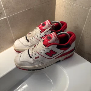 New Balance 550 vita och röda. - Säljer ett par New Balance 550 sneakers i vitt och rött med svarta detaljer. Skorna har klassisk snörning, rund tå och coola basketinspirerade detaljer på plösen. Ovandelen är i skinn och textil, med en chunky sula som ger retrovibbar.