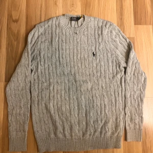 Grå kabelstickad tröja Polo Ralph Lauren - Snygg grå kabelstickad tröja från Polo Ralph Lauren med klassisk rund hals och broderad svart logga på bröstet. Tröjan har ribbade muddar vid ärmslut och nederkant, perfekt för dig som gillar stilren och tidlös design.