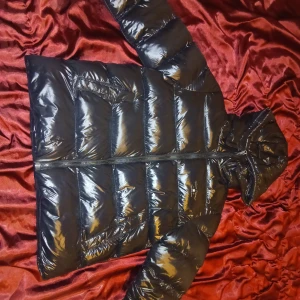 Svart pufferjacka med huva från Vision - Svart, glansig pufferjacka från Vision med huva och dragkedja framtill. Jackan har breda, quiltade sektioner och två sidofickor med dragkedja. Perfekt för kalla dagar och har en modern, sportig look som funkar till streetwear.