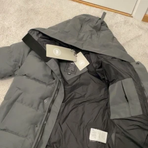 Grå pufferjacka från Canada Goose - Snygg grå pufferjacka från Canada Goose med huva och svart logotyp på ärmen. Jackan har dragkedja och knappar framtill, samt praktiska fickor. Perfekt för kalla dagar med sin tjocka och quiltade design. Stilsäker look och riktigt skön passform.
