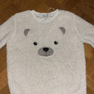 Vit fleece-tröja med björnansikte XS - Supermjuk och fluffig vit fleece-tröja från C i storlek XS. Tröjan har ett gulligt björnansikte broderat framtill med grå detaljer och svarta ögon. Rund hals och långa ärmar med ribbade muddar. Perfekt för dig som gillar mysiga och roliga plagg.