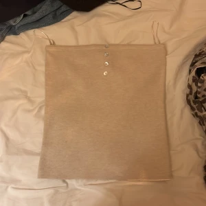 Beige linnetopp från Gina Tricot - Säljer en stilren beige linnetopp från Gina Tricot med smala axelband och fyra dekorativa knappar framtill. Toppen har en rak och enkel form, perfekt för att matcha med jeans eller kjol. Materialet känns mjukt och bekvämt mot huden.
