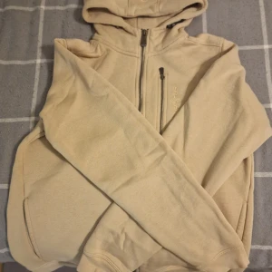 Beige hoodie från Sail Racing med huva - Snygg beige hoodie från Sail Racing med broderad logga på bröstet. Tröjan har huva, hel dragkedja framtill och en extra bröstficka med dragkedja. Tillverkad i mjuk bomullsmix och har långa ärmar. Perfekt för chill dagar och enkel att matcha med jeans.