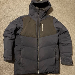 Mörkblå Sail Racing dunjacka M - Säljer en mörkblå dunjacka från Sail Racing i storlek medium. Jackan har svarta paneler på bröst och rygg, huva, två bröstfickor med dragkedja och justerbara ärmslut med kardborre. Perfekt för kalla dagar med sin tjocka och puffiga design. En defekt är vid höger armbåge där det är lite sytt pågrund av ett litet hål innan. 