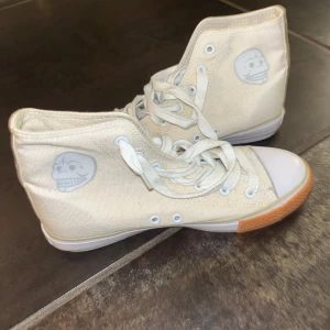 Beige höga sneakers med döskalle - Snygga höga sneakers i beige canvas med vit tå och gummisula i brunt. Skorna har snörning och en cool döskalle-tryck på sidan. Perfekt för dig som gillar unik stil och vill sticka ut lite extra. Klassisk rund tå och platt sula för skön komfort.