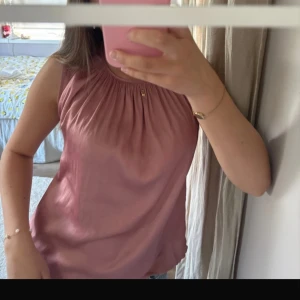 Rosa ärmlös blus med rynkad hals - Säljer en söt rosa blus med rynkad halsringning. Blusen är ärmlös och har en mjuk, flowy passform som känns chill och feminin. Perfekt för varma dagar och enkel att styla med jeans eller kjol.