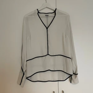 Vit blus med svarta detaljer Gina Tricot - Säljer en vit blus från Gina Tricot i storlek 38. Blusen har svarta kantdetaljer längs v-ringningen, ärmarna och nederdelen, vilket ger en snygg kontrast. Modellen är långärmad och har en loose passform. Perfekt för dig som gillar stilrena och moderna plagg.