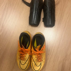  Fotbollsskor och benskydd - Säljer ett par coola fotbollsskor från Nike i gul färg med svarta detaljer och orange snörning. Skorna har en låg profil och är designade för bra grepp på planen. Perfekt för dig som vill sticka ut på matchen! Säljer även ett par benskydd från Puma 