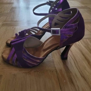 Lila danssandaler med klack - Säljer ett par lila sandaler med klack, perfekta för dans. Skorna har öppen tå, smala remmar i metallic och mocka, samt ett glittrigt spänne vid ankeln. Klacken är smal och ger en elegant siluett. Snygga detaljer och skön passform för dig som gillar att sticka ut. Latin sko (salsa, bachata mm). 9cm klack. Använda bara ett par gånger. Storlek 38.