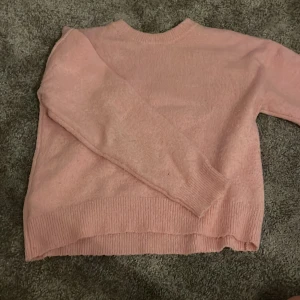 Rosa stickad tröja Gina Tricot - Mysig rosa stickad tröja från Gina Tricot med rund hals och ribbade muddar vid ärmslut och nederkant. Tröjan har en loose passform och långa ärmar, perfekt för att hålla sig varm under kyliga dagar. Enkel och stilren design som är lätt att matcha.