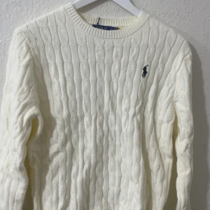 Vit kabelstickad tröja från Polo Ralph Lauren - Säljer en klassisk vit kabelstickad tröja från Polo Ralph Lauren med rund hals och blå broderad logga på bröstet. Tröjan har långärmade och ribbade muddar, perfekt för att hålla värmen under kyliga dagar.