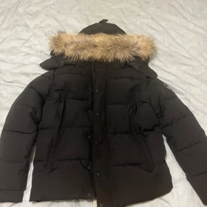 Canada Goose Wyndham Black Badge - Svart pufferjacka från Canada Goose med stor huva och snygg pälsdetalj. Jackan har klassisk logga på ärmen, flera fickor och knappar framtill. Perfekt för kalla vinterdagar och har en riktigt schysst, modern look.