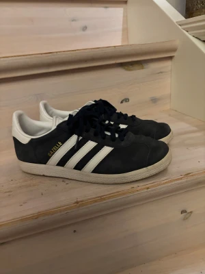 Adidas Gazelle svarta sneakers - Mörk blåa Adidas Gazelle sneakers med klassiska vita ränder och vit häl. Skorna har snörning, platt sula och rund tå. Materialet är mocka med detaljer i syntet. Guldtexten 'GAZELLE' på sidan ger en snygg touch. Perfekt för dig som gillar retrostil och streetwear.