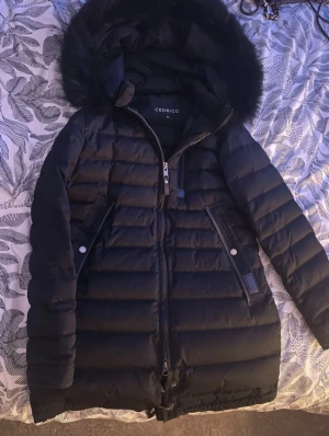 Svart dunjacka från Cedrico med huva - Säljer en svart dunjacka från Cedrico i storlek S. Jackan har en fluffig huva med pälsdetalj, långa ärmar och två fickor med knappstängning. Den är quiltad och har dragkedja framtill. Perfekt för kalla dagar och har en snygg, stilren look.