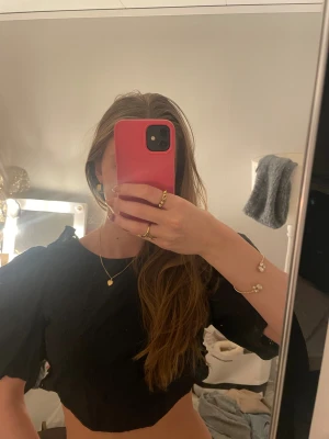 Svart croppad blus med puffärm - Svart blus med croppad passform och puffiga korta ärmar. Toppen har öppen rygg med snörning och knyt baktill. Materialet är lätt och har små broderade hålmönster som ger en cool detalj. Perfekt för dig som gillar statement-ärmar och snygga ryggdetaljer.