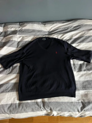 Marinblå v-ringad tröja Polo Ralph Lauren - Svart v-ringad tröja från Polo Ralph Lauren i mjuk pima bomull. Klassisk design med röd broderad logga på bröstet och ribbade muddar vid ärmslut och nederkant. Perfekt för dig som gillar stilrena och tidlösa plagg.