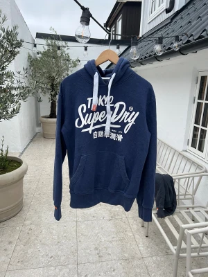 Blå Superdry hoodie med tryck - Säljer en marinblå hoodie från Superdry med stort vitt tryck på bröstet och japanska tecken. Tröjan har huva med vita snören, känguruficka och ribbade muddar. Perfekt för dig som gillar streetwear och snygga loggor.