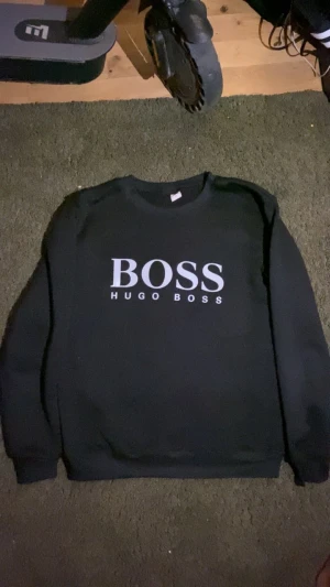 Svart sweatshirt från Hugo Boss - Svart sweatshirt från Hugo Boss med stor vit BOSS-logga på bröstet. Tröjan har rund hals, långa ärmar och en klassisk, enkel design utan extra detaljer. Perfekt för dig som gillar stilrena och snygga märkesplagg. Är M men närmare S