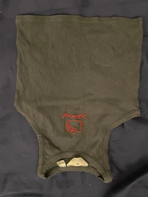 Svart ribbad Hello Kitty tubtopp H&M - Cool svart ribbad tubtopp från H&M med röd Hello Kitty-brodyr framtill. Toppen är ärmlös och har en bandeauformad passning som sitter snyggt. Perfekt för dig som gillar streetstyle och vill sticka ut med en ikonisk detalj.