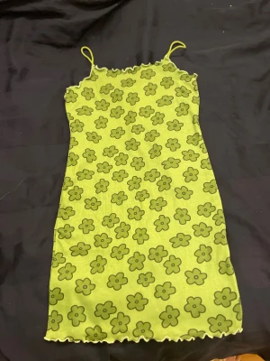 Grön blommig kortklänning med smala axelband - Supersöt grön kortklänning med blommigt mönster och smala axelband. Klänningen har vågig kant upptill och nedtill som ger en extra detalj. Materialet är mjukt och stretchigt, perfekt för sommarens alla dagar. Blommorna är tecknade i mörkare grönt mot en ljusare grön bakgrund.