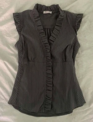 Grå randig blus med volang från Object - Säljer en grå blus från Object med tunna vertikala ränder och volangdetaljer längs krage och ärmslut. Blusen är ärmlös och har en feminin v-ringad omlottfront med volangkant. Perfekt för dig som gillar stilrena detaljer och snygg passform.