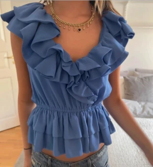 Blå volangblus från H&M - Supersöt blå blus från H&M med volanger längs halsringningen och nedtill. Blusen har en v-ringad design och är ärmlös, med en elastisk midja som ger en snygg siluett. Perfekt för dig som gillar romantiska detaljer och vill sticka ut med färg. Priset går att diskutera😊