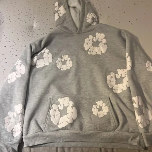 Grå hoodie med blommor från Denim Tears - Säljer en grå hoodie från Denim Tears med stora vita blommor tryckta över hela tröjan. Tröjan har huva, känguruficka och är långärmad. Perfekt för dig som vill sticka ut med en unik och trendig stil.