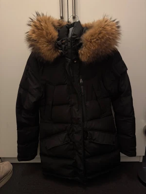 Svart dunjacka med päls från Hollies - Säljer en svart dunjacka från Hollies med fluffig brun päls runt huvan. Jackan har quiltad design, dragkedja och knappar framtill, samt praktiska fickor med lock. Perfekt för kalla vinterdagar och har en snygg, normal passform. Är som ny skick