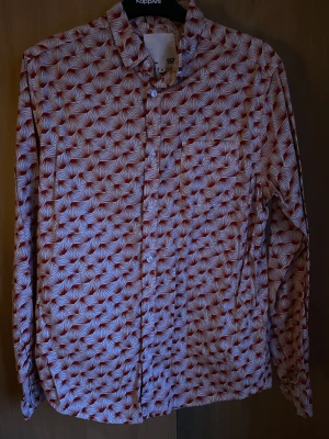 Rödvit mönstrad skjorta från Lager157 - Snygg skjorta från Lager157 med ett unikt rött och vitt mönster som påminner om blad eller grafiska former. Skjortan har klassisk krage, långa ärmar och knappar framtill. Perfekt för dig som vill sticka ut med en cool och färgstark look.